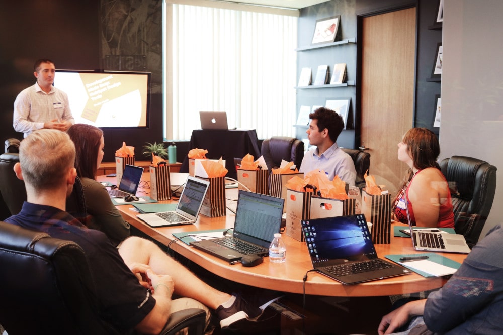 Man presenting to a group of people sitting around a table with laptops and gift bags